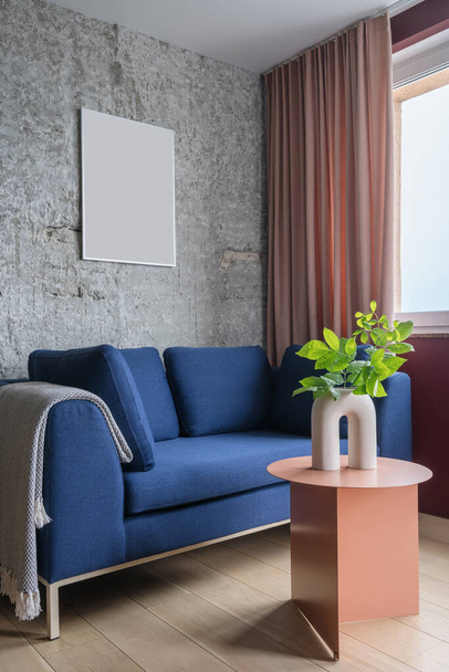 プレイやクッション付きの青い快適なソファ付きのスタイリッシュなアパート、サイドテーブルの植物とコンクリート灰色の壁にコピースペースとポスター付きのセラミック花瓶 - 写真・画像