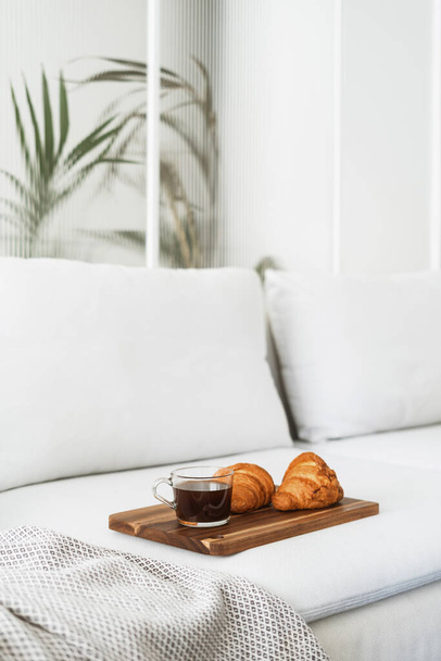 breakfast at home, coffee drink in glass cup and sweet french croissant on wooden board on comfort couch at apartment with light interior - Zdjęcie, obraz