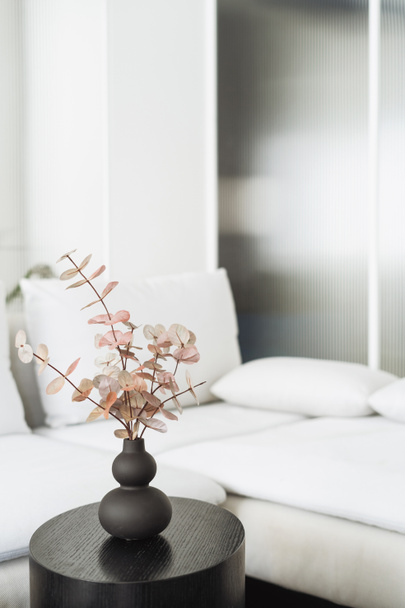 rama de eucalipto en jarrón de cerámica sobre mesa auxiliar redonda de madera y sofá con almohadas sobre fondo en apartamento con interior escandinavo - Foto, imagen