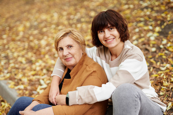 Portrait of old pensioner mom and middle aged cheerful daughter spending time together outdoor at falling season. Caucasian senior mother and her daughter sitting in adorable autumn park - Photo, Image