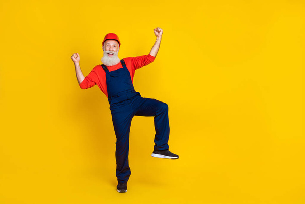 Photo of positive funny senior guy dressed uniform overall red hardhat rising fists empty space isolated yellow color background. - Fotó, kép