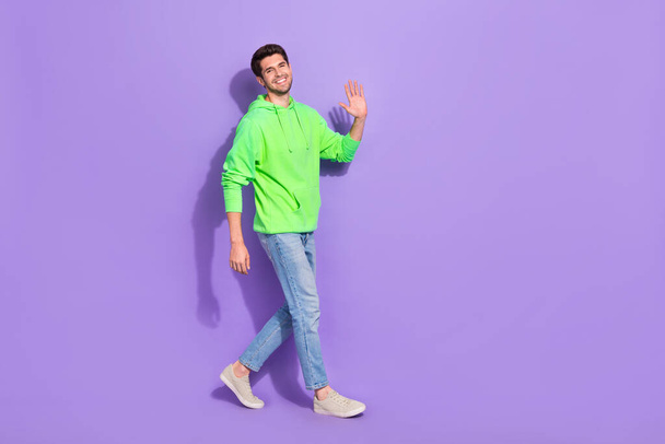 Full length photo of cheerful cute man wear green hoodie walking waving arm empty space isolated purple color background. - Φωτογραφία, εικόνα