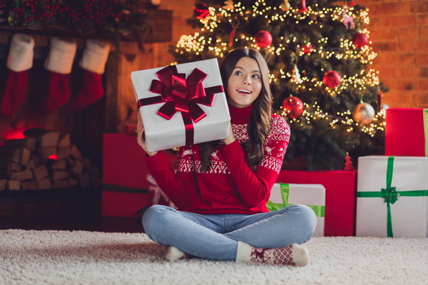 Photo of cheerful good mood nice girl with curly hairstyle wear red ornament jumper holding present look empty space in house indoors. - Foto, imagen