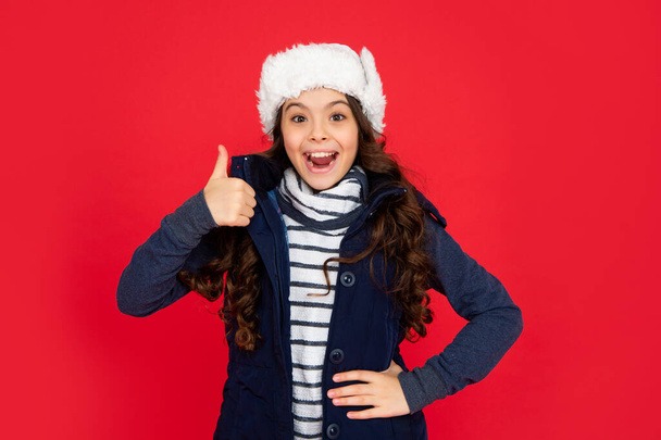 winter fashion. thumb up. surprised kid with curly hair in earflap hat. teen girl on red background. portrait of child wearing warm clothes. express positive emotion. - Fotoğraf, Görsel