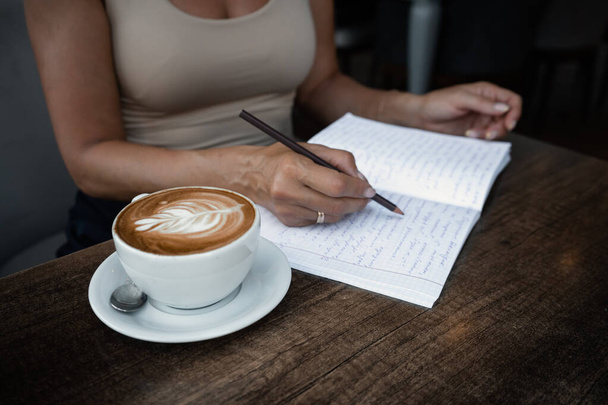 Mujer irreconocible se sienta a la mesa en la cafetería con una taza de café y escribe en notas de cuaderno escribiendo información. Foto de alta calidad - Foto, Imagen