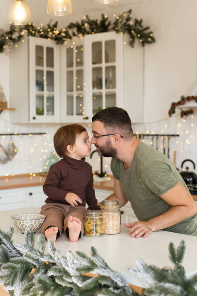 Papá e hijo jugando y divirtiéndose en la cocina decorada de Navidad. Feliz familia esperando las fiestas. Imagen vertical - Foto, imagen
