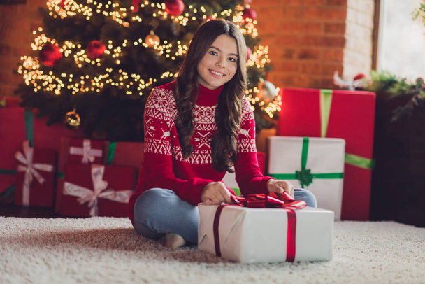 Portret dobrego nastroju zadowolona wdzięczna dziewczyna z falistą fryzurę ubrany czerwony sweter ozdoba rozpakowywanie prezent pudełko w domu wewnątrz. - Zdjęcie, obraz