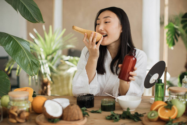 Beautiful asian woman preparing body milk or cream from pumpkin for skin care sitting at wooden table with various ingredients for making natural cosmetics on light exotic studio background. - Photo, Image