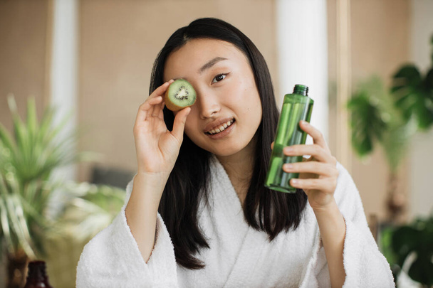 Kiwi tonic oil skin and hair care, home spa concept. Closeup portrait of beautiful asian girl making nourishing mask with kiwi in tropical exotic studio. - Photo, image