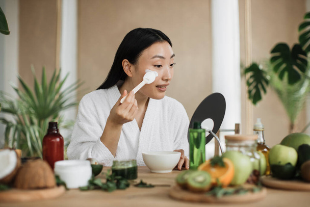 Healthy asian woman sitting at wooden table with various ingredients preparing natural cosmetics at home mixing mass in bowl while preparing cream and applying on face using wooden spoon. - Photo, image