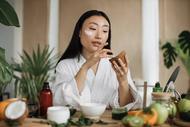 Blogger or content creator asian woman preparing applying natural cosmetics at home, holding coconut for homemade cream for skin, hair care sitting at wooden table with exotic background in home spa - Zdjęcie, obraz