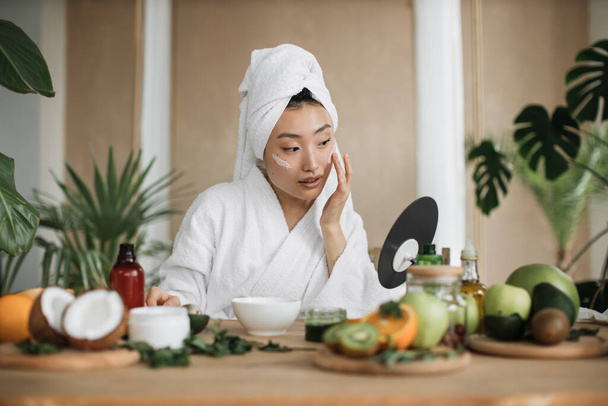 Healthy asian woman in white bathrobe and towel sitting at wooden table with various ingredients preparing natural cosmetics at home mixing mass in bowl while preparing cream and applying on face. - 写真・画像