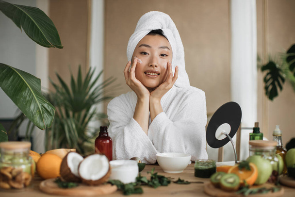 Close up view of face of young asian woman applying natural cosmetics at home, making homemade cream for skin, hair care at with exotic background in spa resort - Fotografie, Obrázek