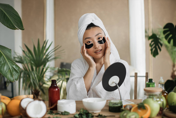 Asian woman sitting at table in white towel and bathrobe, looking at mirror and applying patches under eyes. Beautiful female taking care of her facial skin in exotic background. - Foto, Imagen