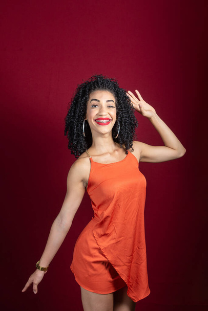 Young beautiful smiling woman, standing, smiling, wearing orange colored outfit against red background in studio. - Photo, Image