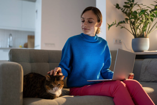 Relaxed Italian young woman student or freelancer sitting on cozy couch holding laptop on lap working studying from home strokes with fluffy pet cat in living room with plants. Togetherness concept. - Photo, Image