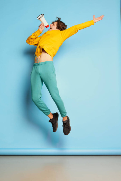 Portrait of young man with curly hair posing, shouting in megaphone in a jump isolated over blue background. Concept of youth culture, emotions, facial expression, fashion. Copy space for ad - Foto, Imagen