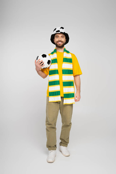 full length of happy bearded sports fan in hat holding football on grey - Photo, Image