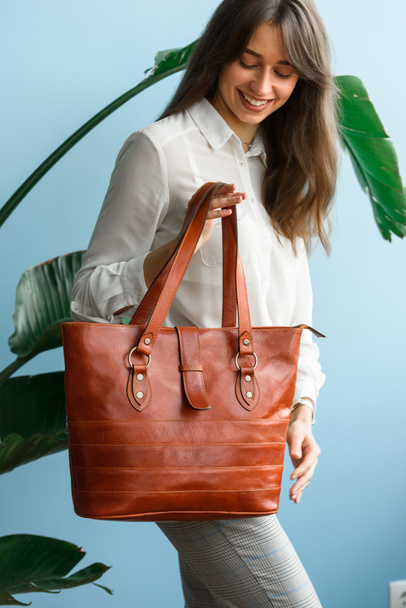 close-up photo of orange leather bag in a womans hands. indoor photo. beautiful slender girl in gray plaid pants and a white blouse posing with a handbag - Fotografie, Obrázek