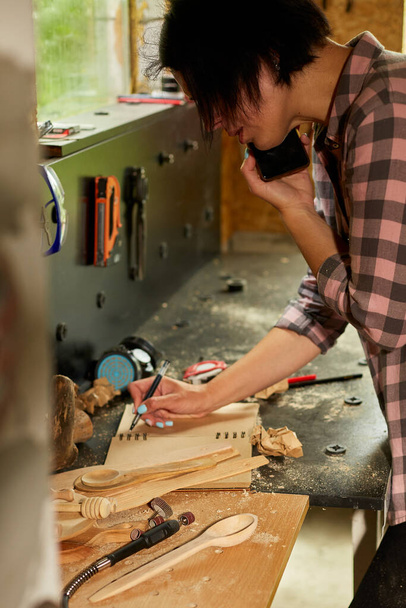 Cheerful carpenter woman discussing order details with her customer on smartphone and makes notes at workshop, Creating craft handmade souvenirs, female in workshop. - 写真・画像