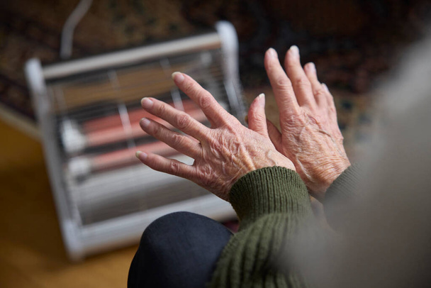 Close Up Of Senior Woman Warming Hands On Electric Heater In Cosyt Of Living Energy Crisis - Zdjęcie, obraz