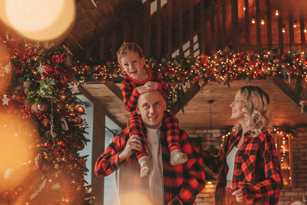 Uśmiechnij się aktywnych rodziców z małym synem w czerwonym kratkę śpiochy czeka Santa kryty. Stylowa rodzina uwielbia się przytulać do domu. Świętuj Nowy Rok girlandy i światła w wigilię 25 grudnia Noel tree - Zdjęcie, obraz