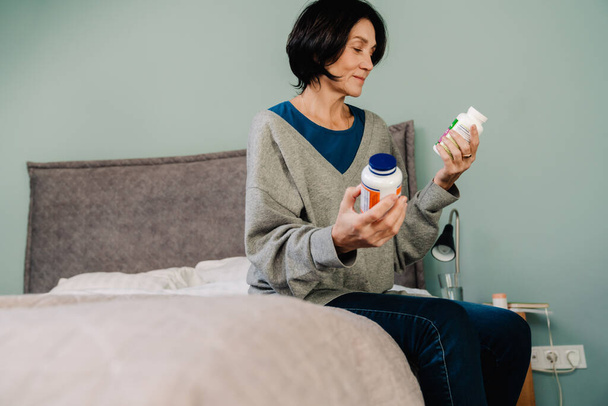 White mature woman examining medicine while sitting on bed at home - Photo, Image