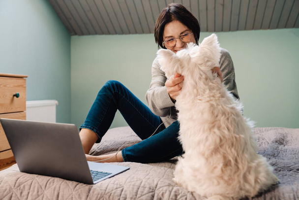 White mature woman using laptop while petting her dog on bed at home - Foto, Bild