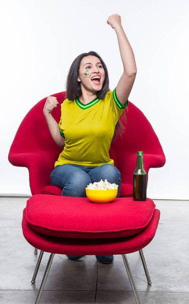 Beautiful young woman fan in a yellow t-shirt and flag painted on face of the Brazil national team celebrating happy sitting in a red armchair with a sandwich and beer on a stool. - Foto, imagen