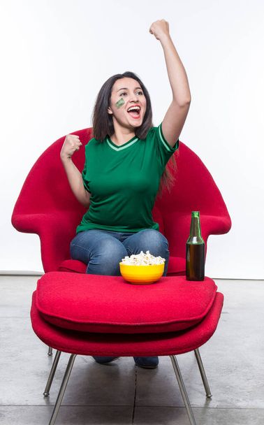 Beautiful young woman fan in a green t-shirt and flag painted on face of the Saudi Arabia national team celebrating happy sitting in a red armchair with a sandwich and beer on a stool. - Foto, immagini