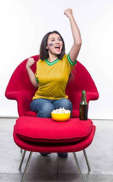 Beautiful young woman fan in a yellow t-shirt and flag painted on face of the Australia national team celebrating happy sitting in a red armchair with a sandwich and beer on a stool. - Zdjęcie, obraz