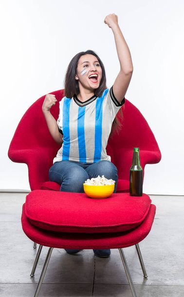 Beautiful young woman fan in a light blue and white t-shirt and flag painted on face of the Argentina national team celebrating happy sitting in a red armchair with a sandwich and beer on a stool. - Foto, Imagem
