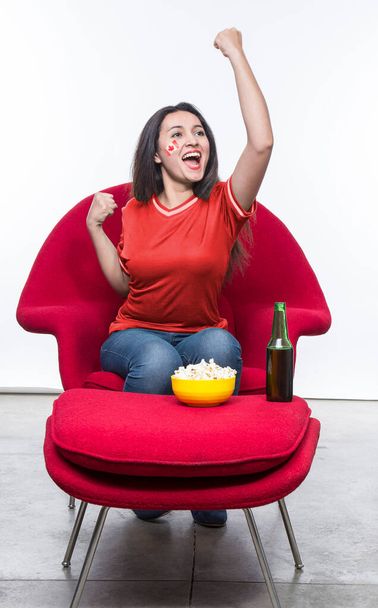 Beautiful young woman fan in a red t-shirt and flag painted on face of the Canada national team celebrating happy sitting in a red armchair with a sandwich and beer on a stool. - Foto, Bild