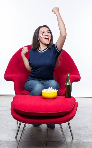 Beautiful young woman fan in a blue t-shirt and flag painted on face of the France national team celebrating happy sitting in a red armchair with a sandwich and beer on a stool. - Foto, Bild