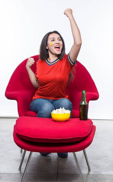 Beautiful young woman fan in a red t-shirt and flag painted on face of the Spain national team celebrating happy sitting in a red armchair with a sandwich and beer on a stool. - Foto, imagen