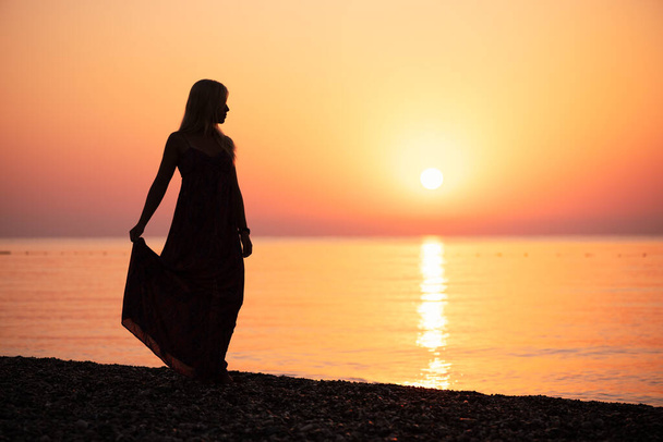 A woman in a sundress contemplates a vivid sunset by the sea - Photo, Image
