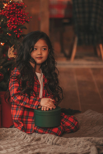 Šťastné Japonsko malá dívka snílek čeká zázrak od Santa pózování na podlaze poblíž stromu Noel. Asijské dítě v červené kostkované oblečení slaví nový rok s dárky krabice na bokeh vánoční světla předvečer 25 prosince - Fotografie, Obrázek