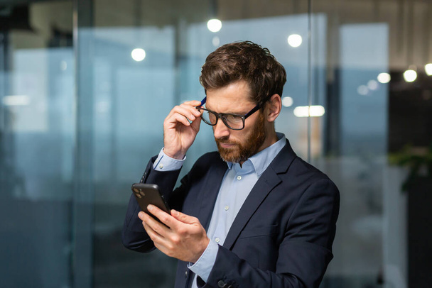 Mature businessman in office near window in evening upset reading message online notification from phone, male boss working inside office in business suit and glasses with beard, upset investor. - Foto, Imagem