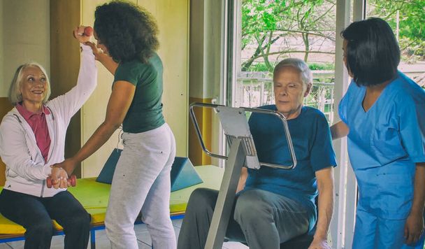 Concepto de rehabilitación. Ancianos jubilados pareja entrenamiento en un gimnasio supervisado por experto africano y asiático mujeres entrenadores. - Foto, Imagen