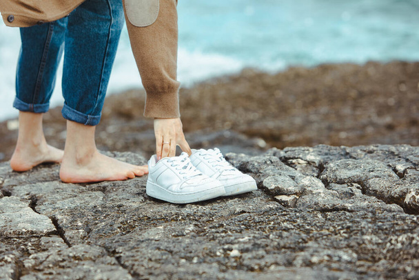 γυναίκα περπατώντας ξυπόλητη δίπλα στη θάλασσα κρατώντας τα χέρια άσπρα παπούτσια. καλοκαιρινές διακοπές. Ένωση φύσης - Φωτογραφία, εικόνα