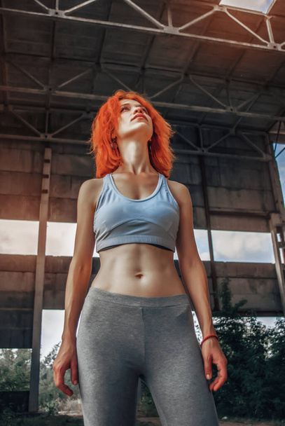 Strong female athlete wearing sportswear taking break after exercising. Fit woman standing inside abandoned warehouse. - Photo, Image