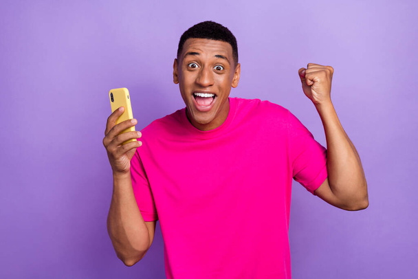 Photo of impressed successful lucky guy box haircut pink t-shirt hold phone clench fists win bet isolated on purple color background. - Foto, Imagen