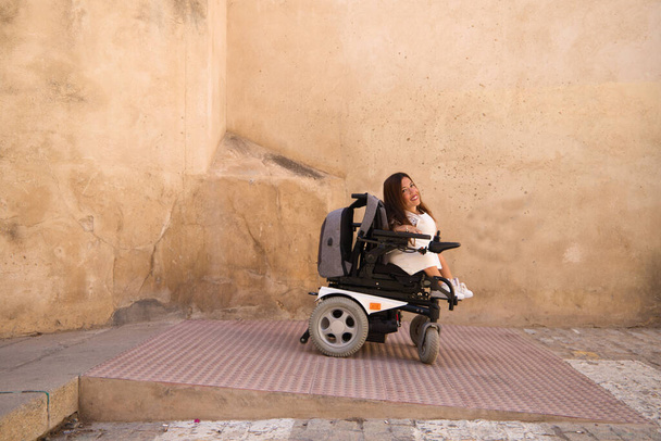 schöne junge behinderte Frau im Rollstuhl mit eingeschränkter Mobilität fährt einen für Rollstuhlfahrer zugänglichen Hang hinunter. Konzept zur Beseitigung architektonischer Barrieren - Foto, Bild