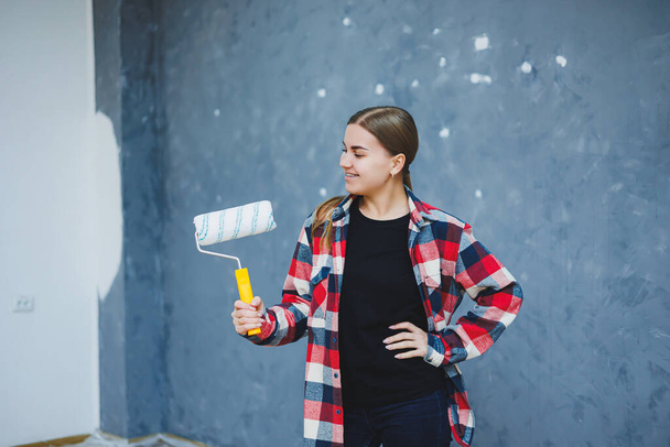 A beautiful young woman in a checkered shirt is renovating her room, holding a paint roller - Foto, imagen