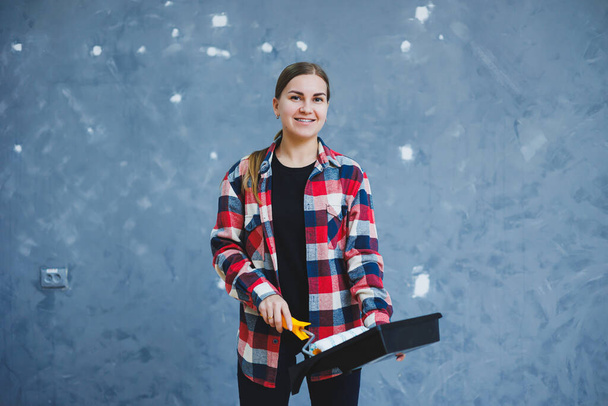 Una hermosa joven con una camisa a cuadros está renovando su habitación, sosteniendo un rodillo de pintura - Foto, imagen