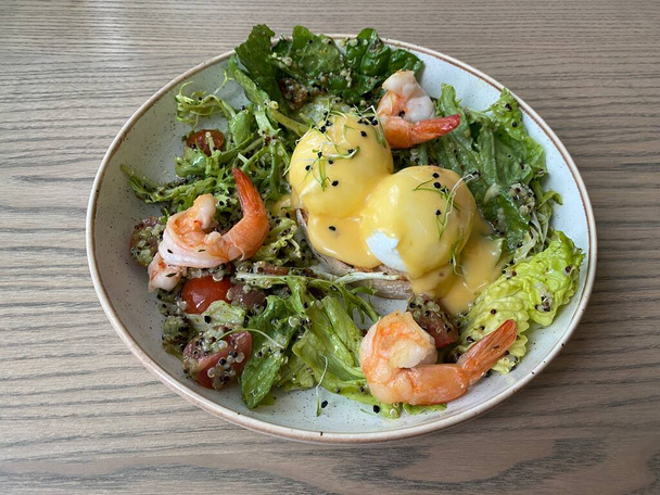 Café da manhã saudável LCHF. Ovos Benedict com quinoa, salada fresca, camarão e molho em um prato bege em uma mesa de madeira - Foto, Imagem