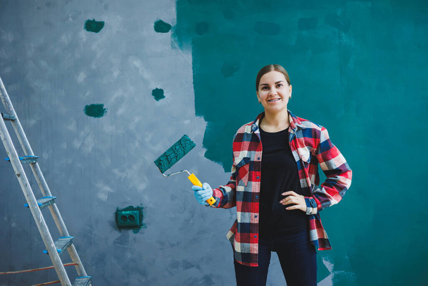 若い美しい女性デザイナーは、修理を行い、ローラーで緑の塗料で灰色の壁をペイントします。再生シャツ、長い髪とジーンズ. - 写真・画像
