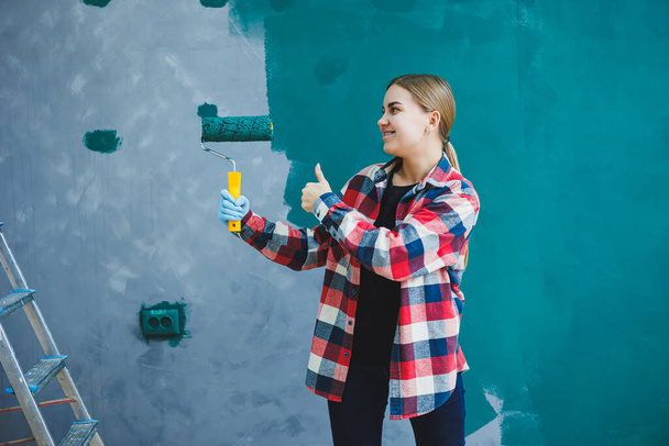 A young beautiful woman designer makes repairs, paints a gray wall with green paint with a roller. Plaid shirt, long hair and jeans. - Valokuva, kuva