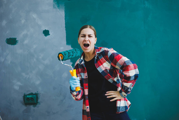 An emotional woman paints the walls. Young smiling pretty woman in shirt holding roller in right hand and singing. Repair in the room. - Foto, afbeelding