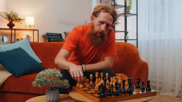 Focused caucasian bearded man chessman sitting at home room couch learning playing chess board game alone, educational leisure domestic activity, strategic skills. Young guy at home on orange couch - Photo, Image